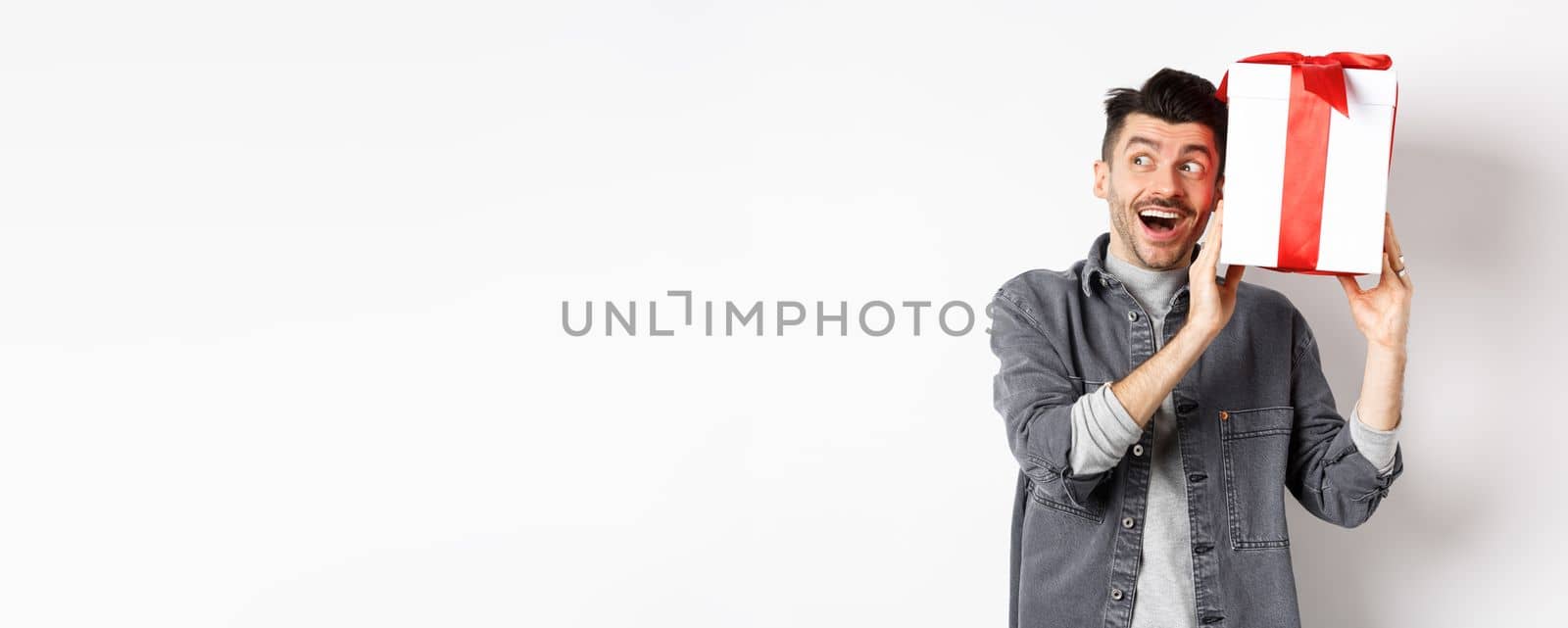 Happy boyfriend shaking his gift to guess what inside, receive surprise present on Valentines day, smiling and looking aside cheerful, standing on white background by Benzoix