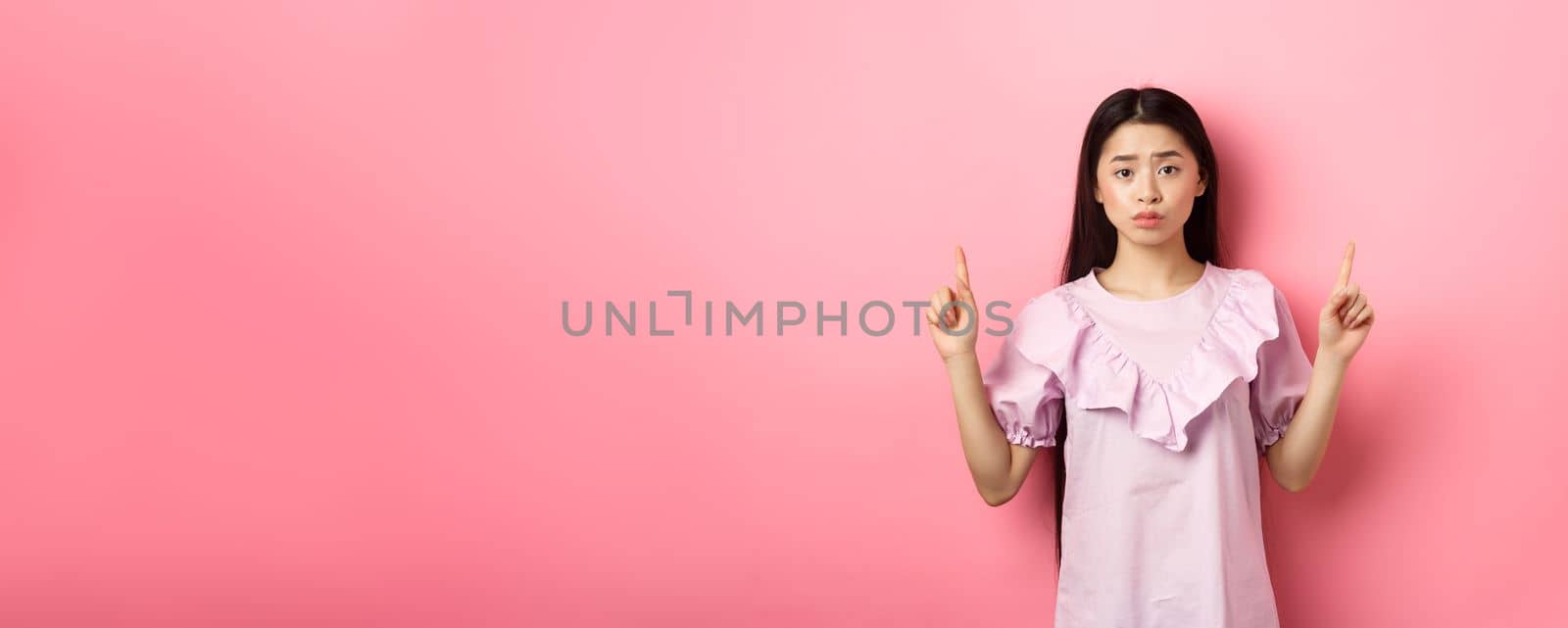 Cute timid asian woman pointing fingers up, frowning and look upset, pointing fingers up at logo, standing in dress on pink background.