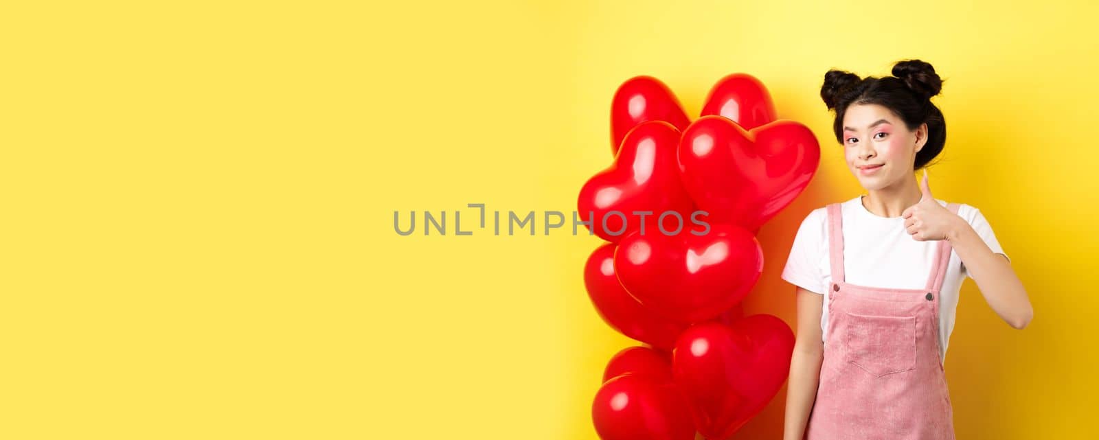 Fashionble asian woman in romantic outfit with make-up, showing thumb up and smiling, praising Valentines day offer, standing near red heart balloons, yellow background.