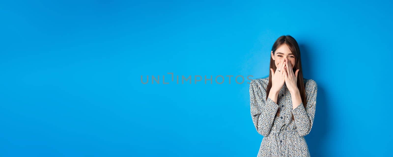 Cheerful caucasina girl in dress laughing and having fun, covering mouth with hands and chuckle over something funny, standing on blue background.