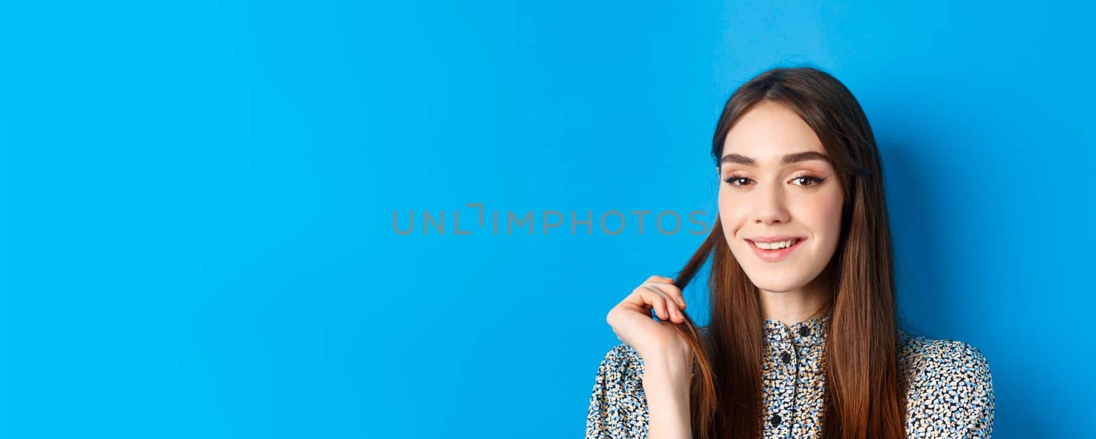 Close-up portrait of flirty young woman with natural beauty, smiling and playing with hair strand, having interesting idea, standing on blue background.