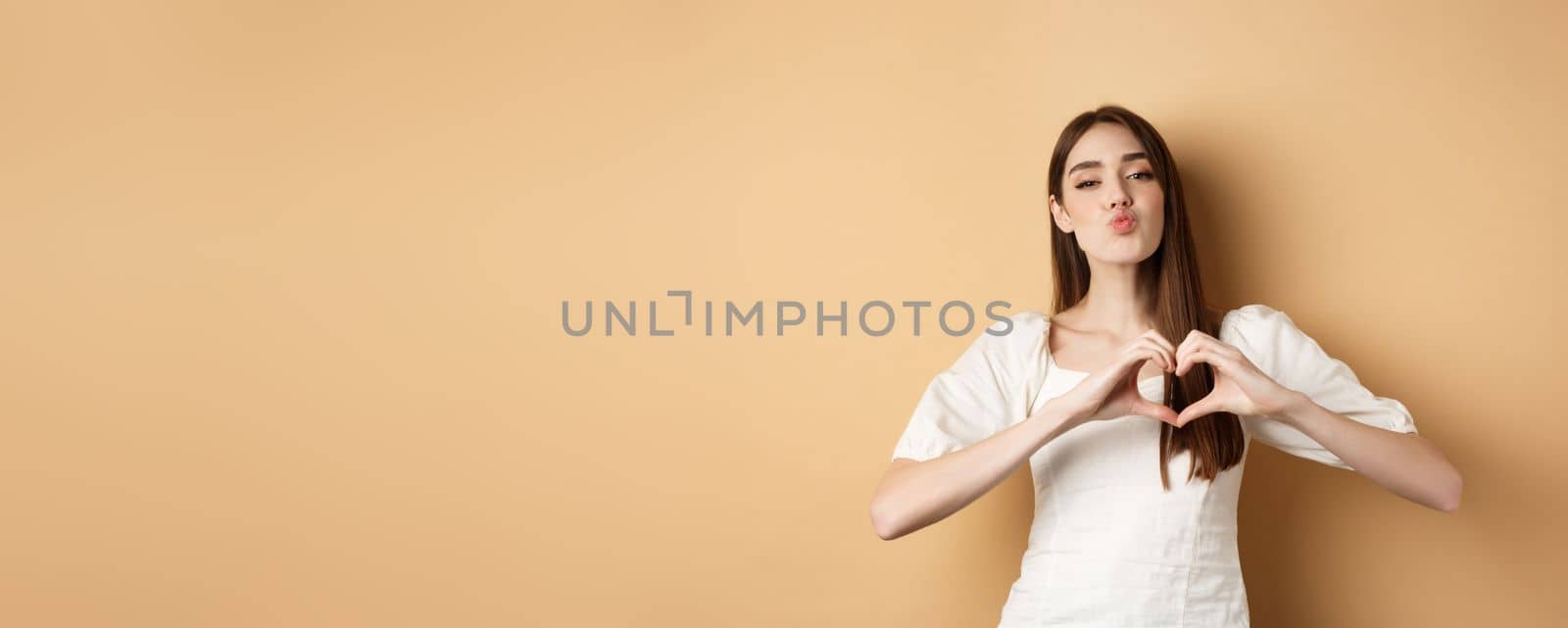 Valentines day. Lovely young womam in white dress, confess in love, pucker lips for kiss and showing heart gesture to lover, standing on beige background.