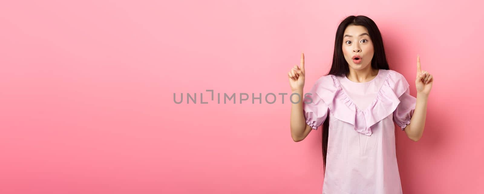 Excited asian girl showing awesome promo offer, pointing fingers up and gasping amazed, standing on pink background by Benzoix
