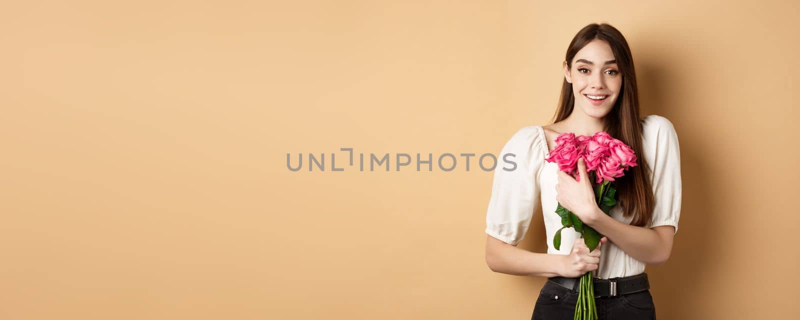 Valentines day. Romantic girl smiling happy at camera, holding bouquet of pink roses from lover, standing on beige background.