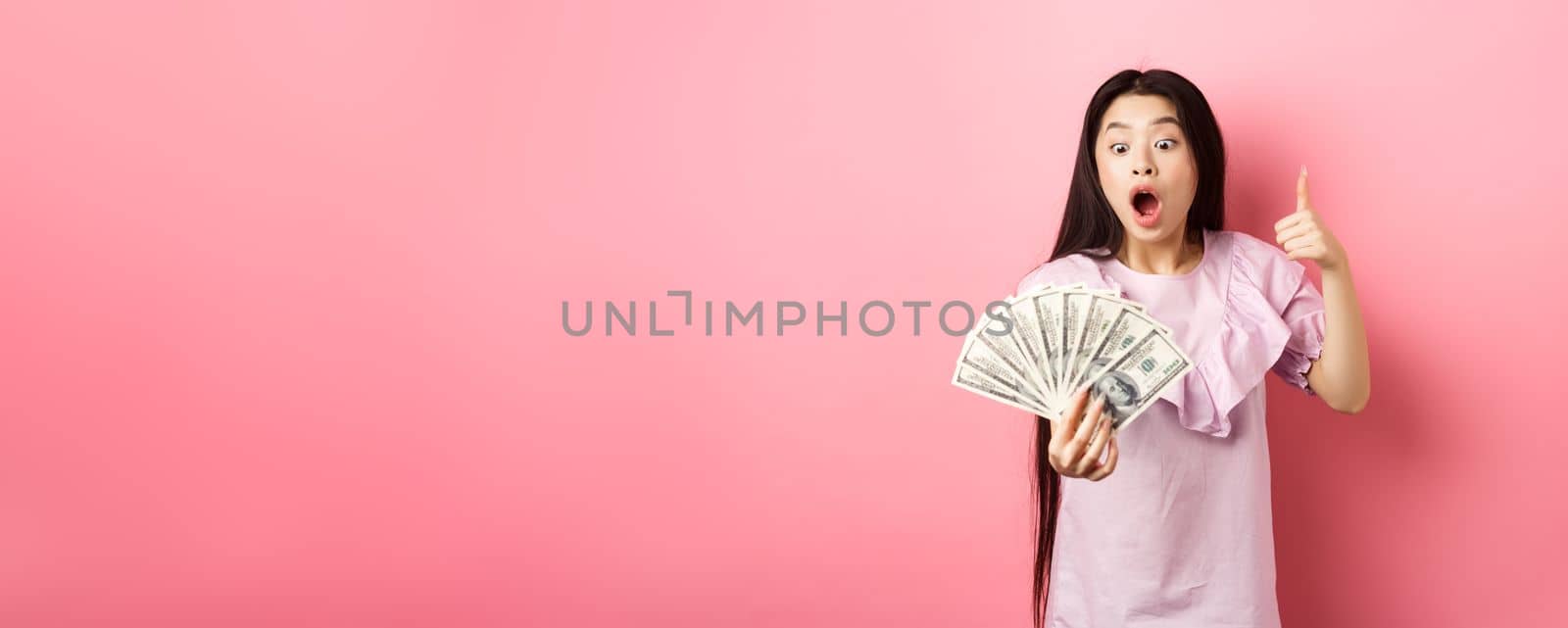 Excited teen girl holding big amount of money, showing dollar bills and thumbs up, standing amazed on pink background by Benzoix