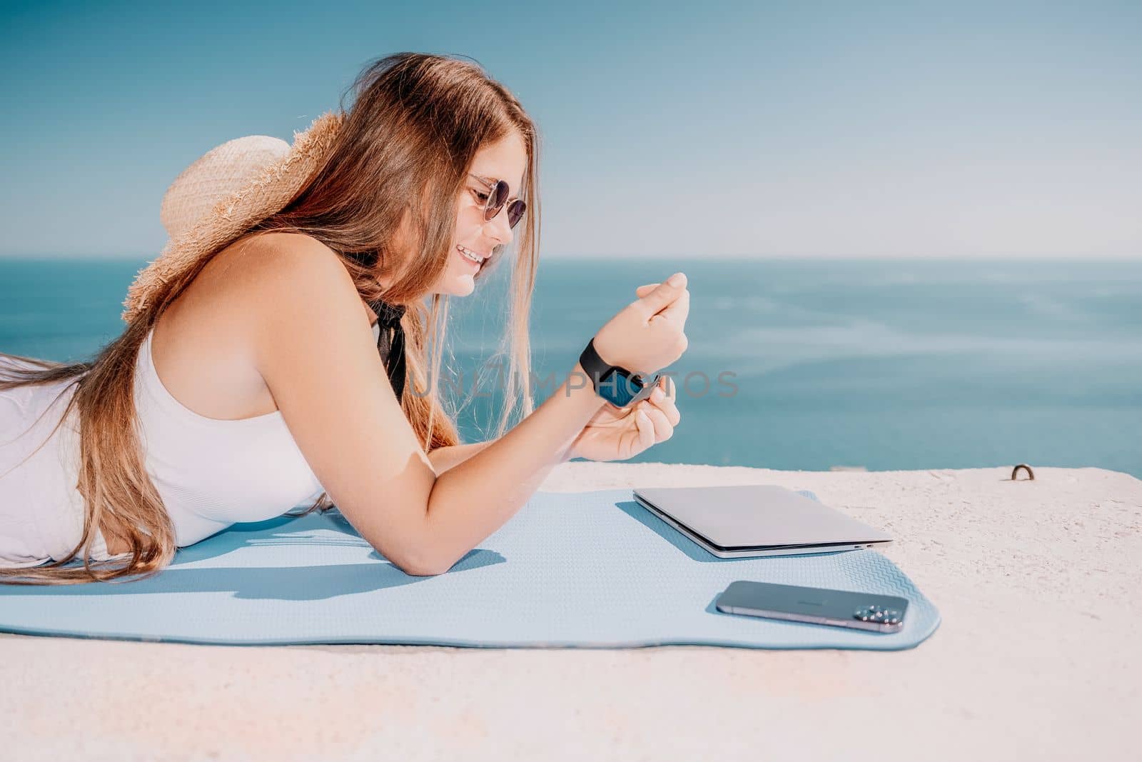 Successful business woman in yellow hat working on laptop by the sea. Pretty lady typing on computer at summer day outdoors. Freelance, travel and holidays concept.