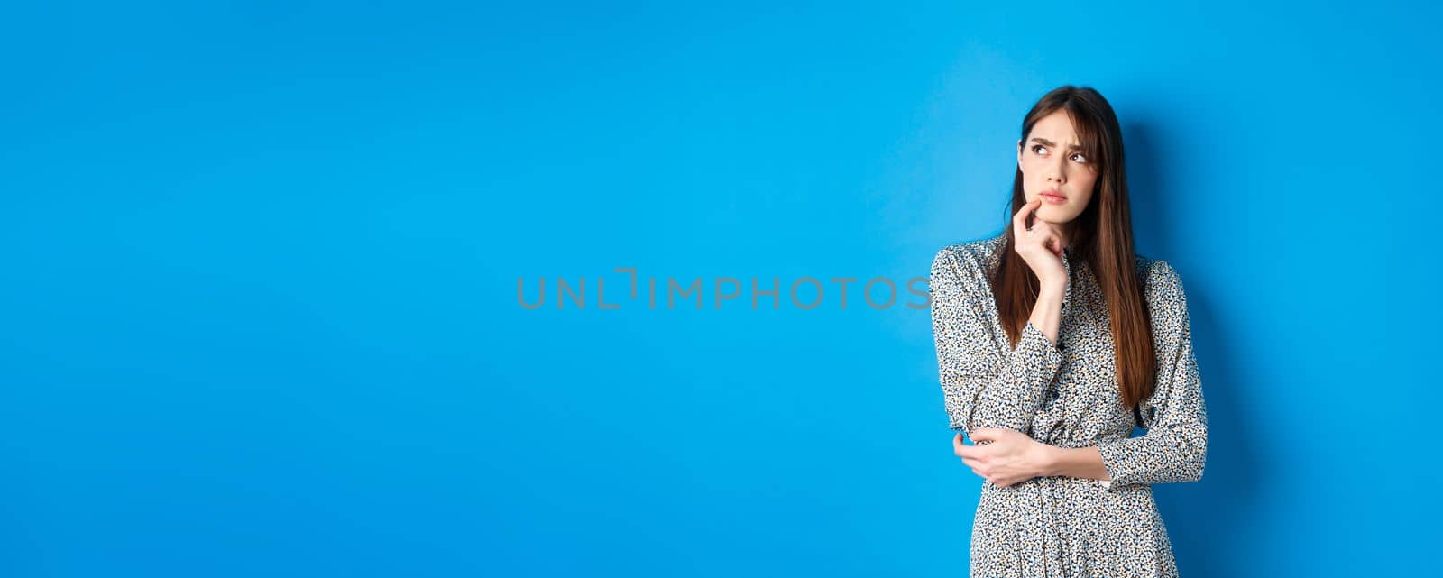 Thoughtful serious woman in dress looking away pensive, standing hesitant and unsure, thinking on blue background.