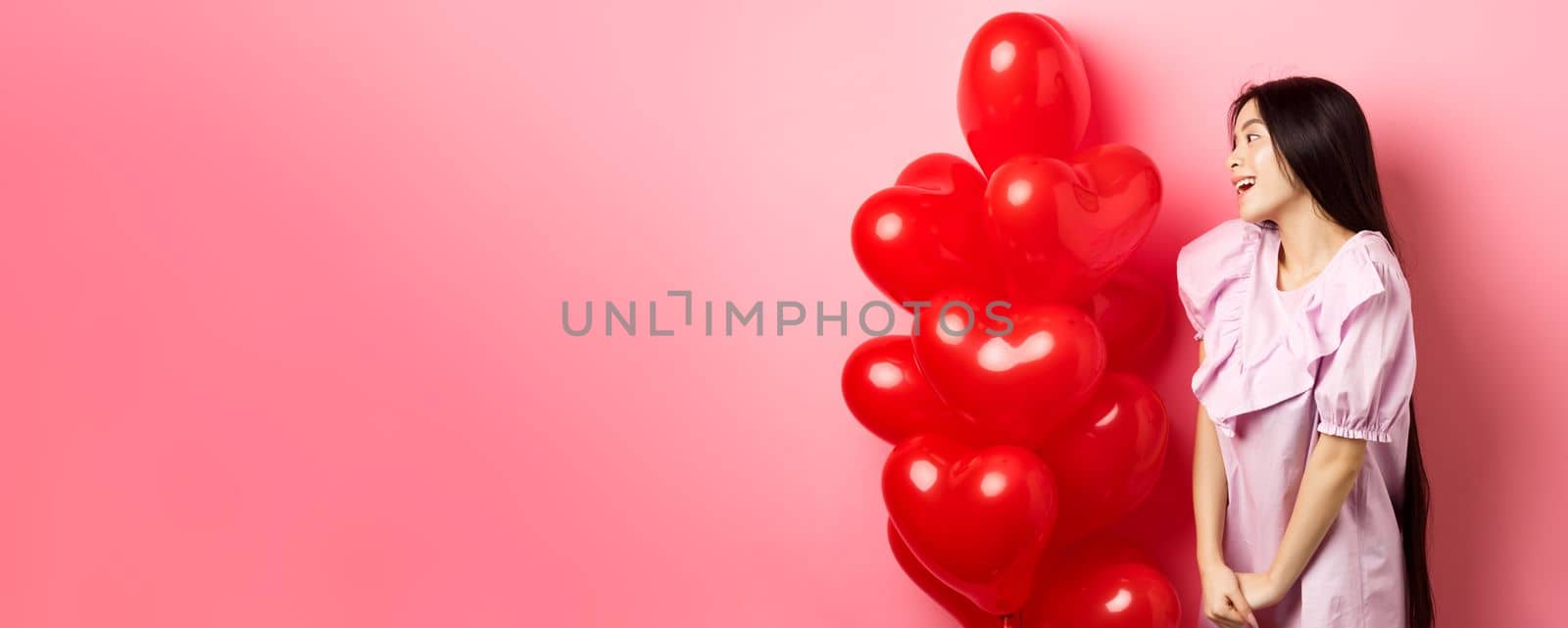 Romance and love concept. Romantic asian girl looking with affection and sympathy at empty space, standing near red hearts balloons from lover, pink background.