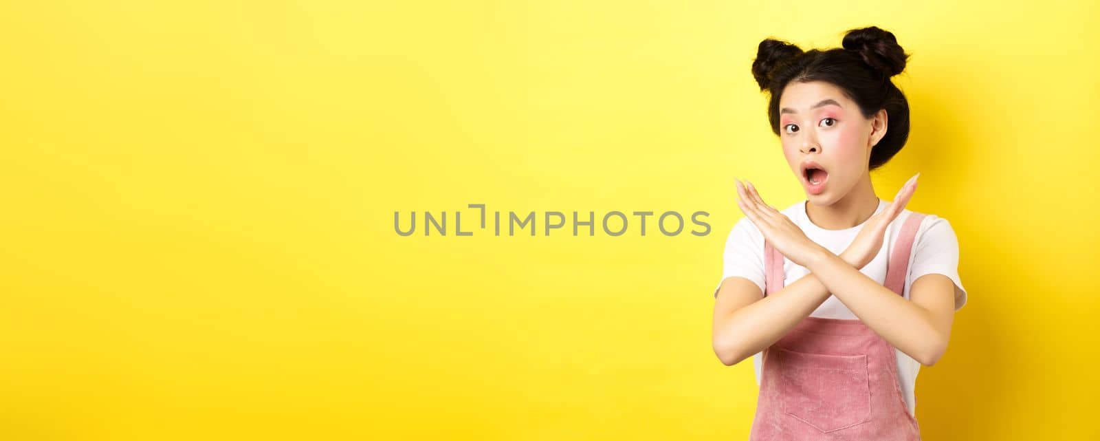 She says no. Asian beauty girl with makeup, showing cross sign, stop and reject offer, standing disappointed on yellow background.