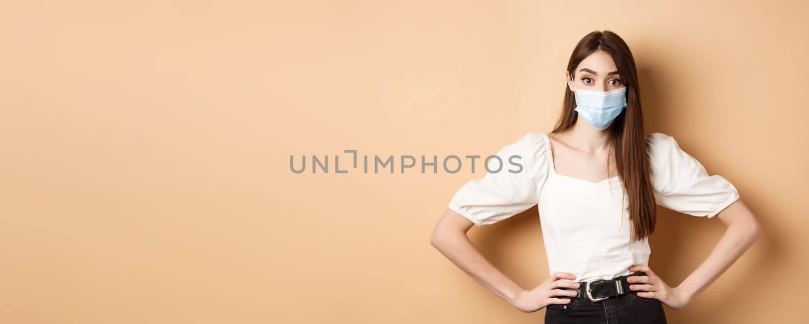 Covid-19 and lifestyle concept. Woman in medical mask looking alarmed at camera, standing on beige background by Benzoix