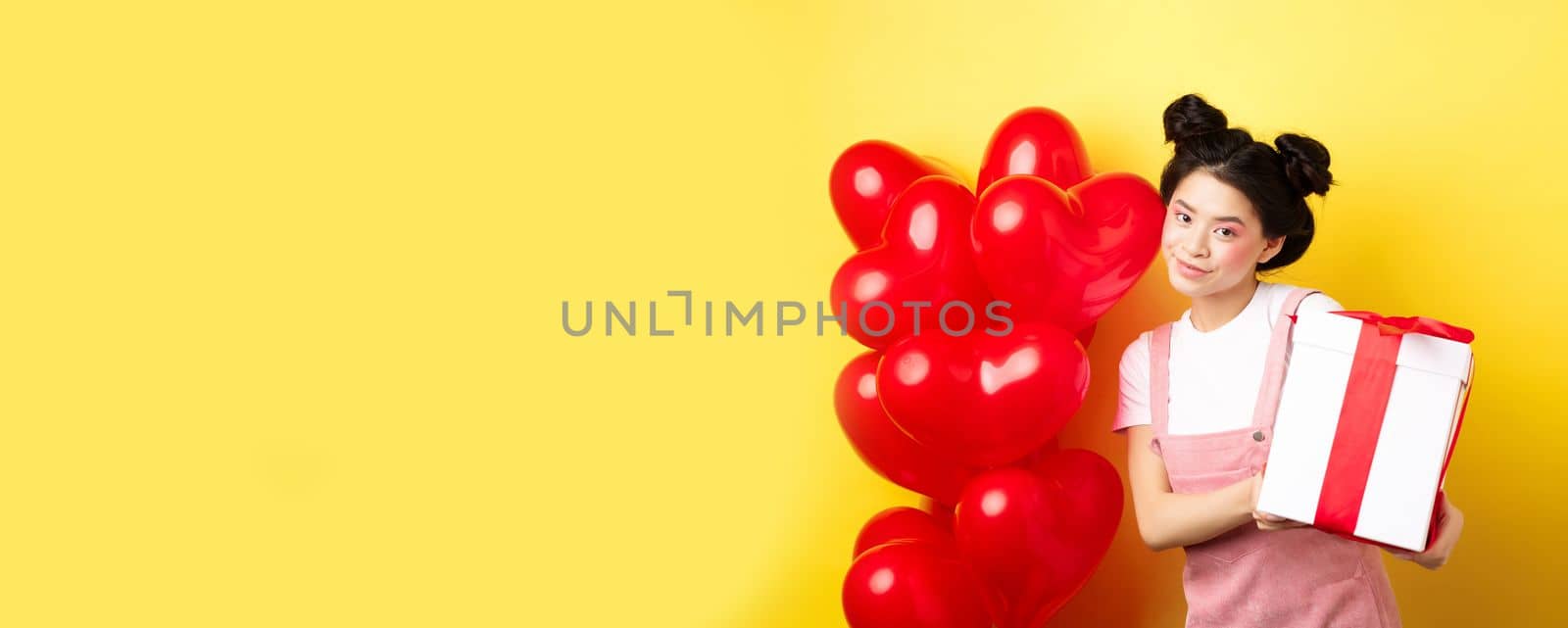 Happy Valentines day. Beautiful and stylish asian woman posing near heart balloons with lovely box with gift from lover, standing over yellow background by Benzoix