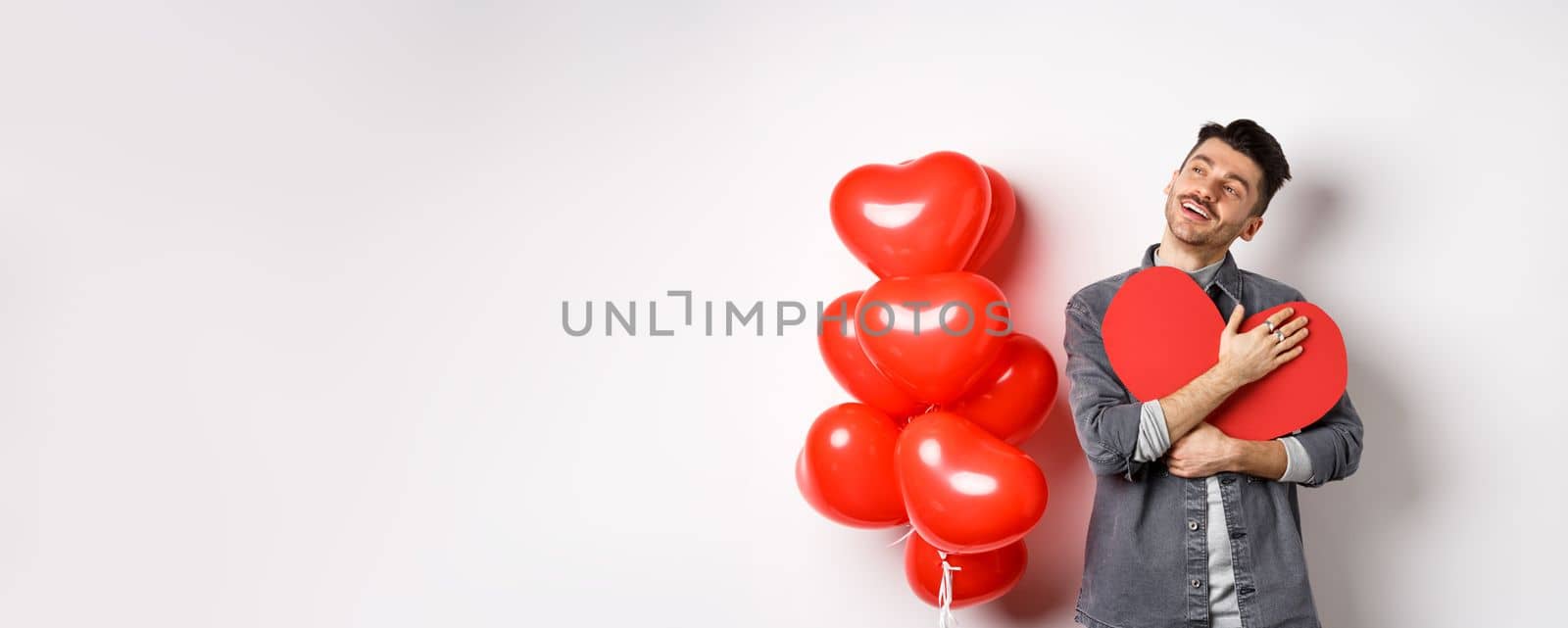 Romantic man hugging big red heart cutout and looking away dreamy, thinking of girlfriend and valentines day, imaging lover, standing on white background.