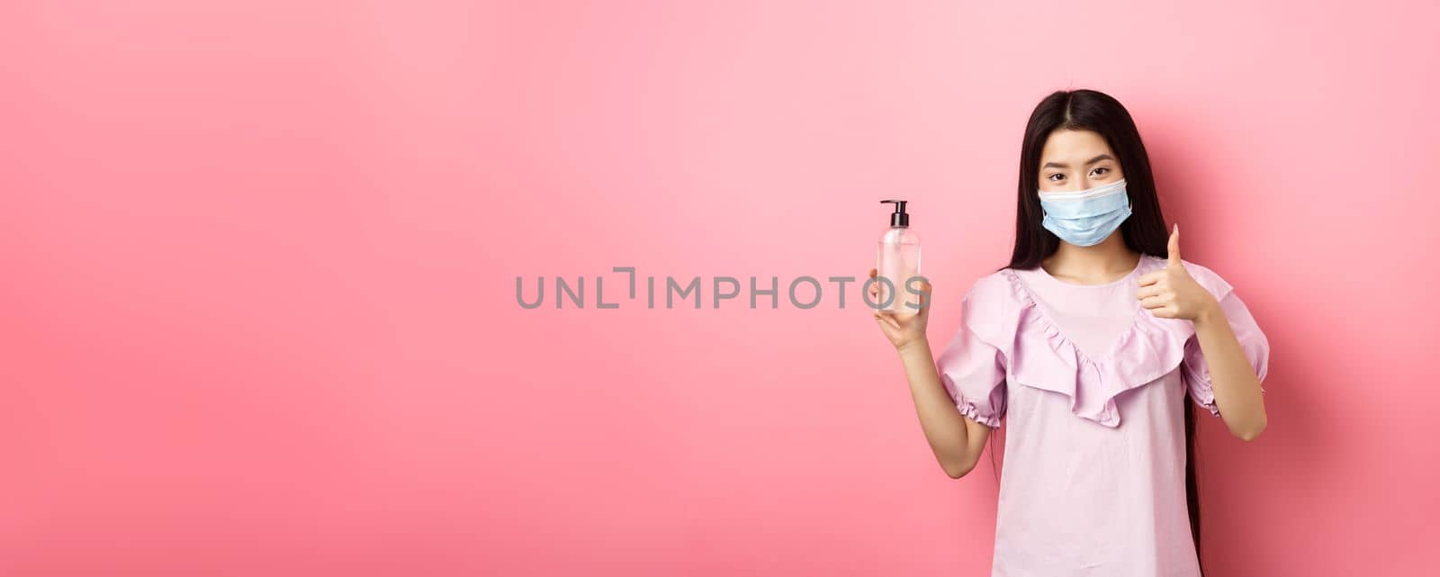 Healthy people and covid-19 pandemic concept. Cheerful asian woman in medical mask recommend hand sanitizer, showing thumb-up, praise antiseptic bottle, white background.