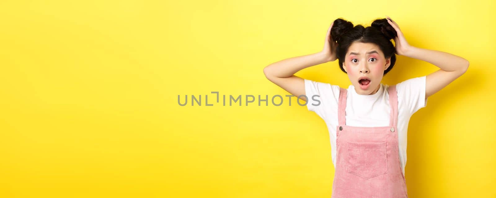 Shocked asian girl gasping worried, checking her hairstyle, standing anxious on yellow background.