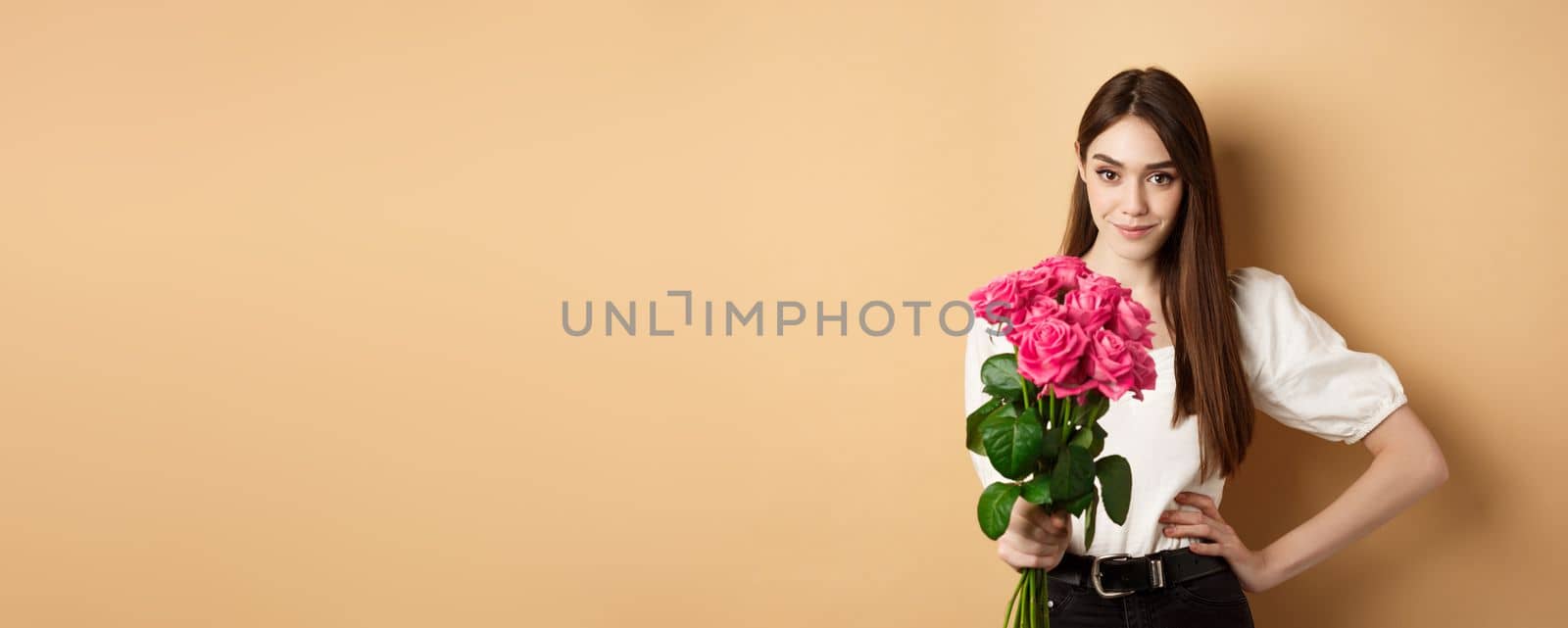 Valentines day. Beautiful girlfriend holding pink roses and looking at camera. Young woman receive flowers from her date, standing on beige background.