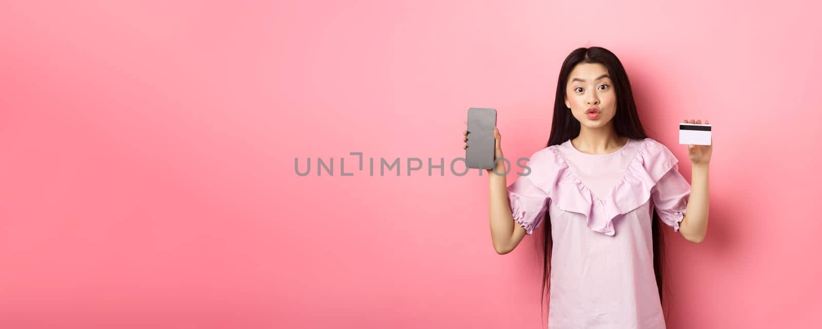 Online shopping. Excited asian woman showing plastic credit card with empty smartphone screen, advertising internet shop, standing on pink background.