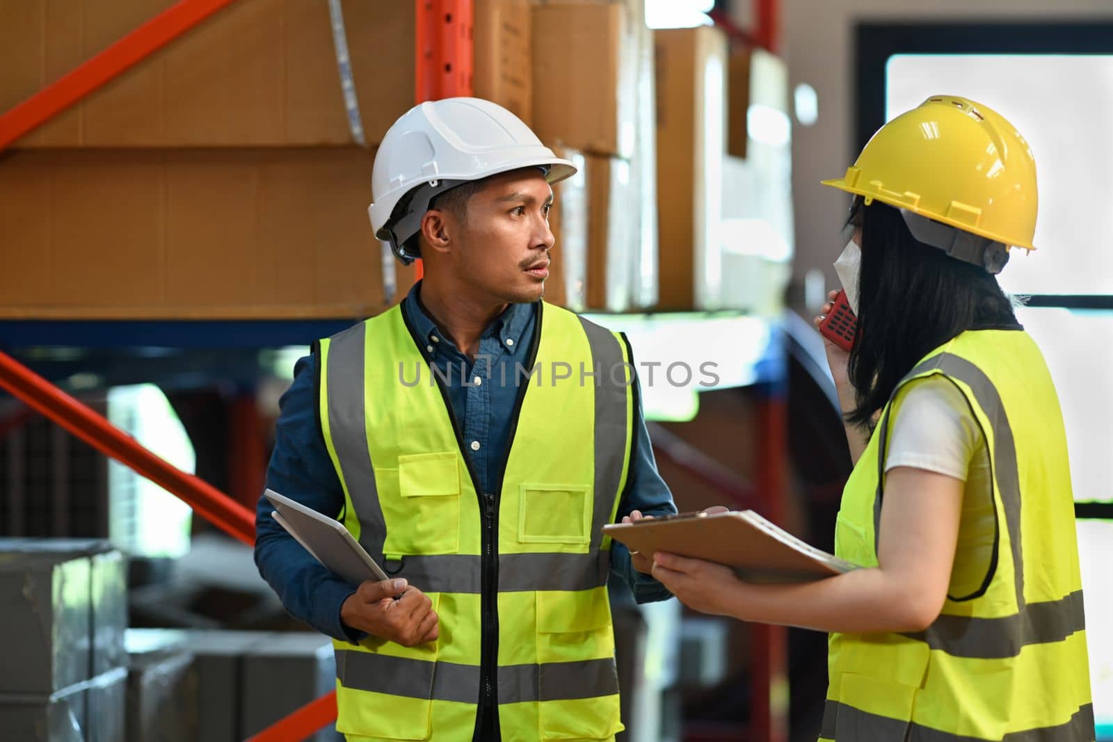Two warehouse coworkers talking to each other while standing between rows of tall shelves full of packed boxes and goods.