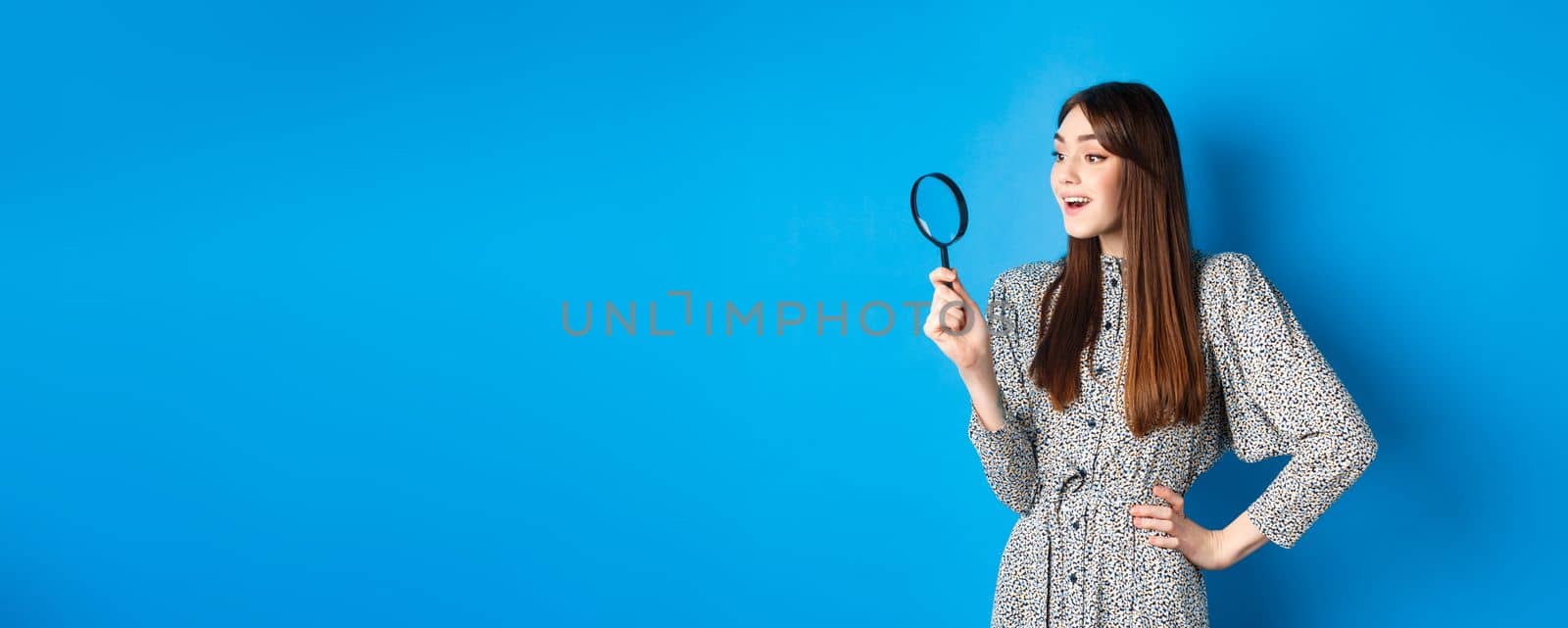 Excited girl looking left with magnifying glass, found interesting promo, investigating or searching, standing on blue background.