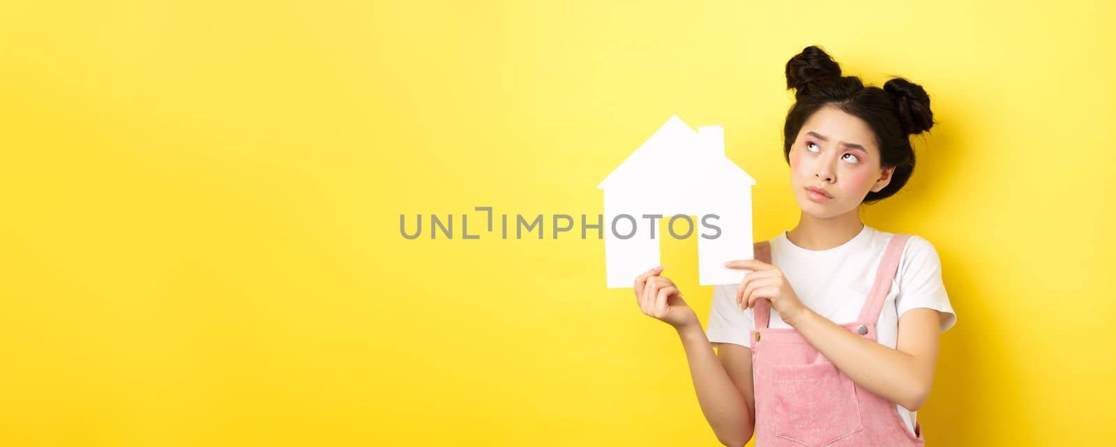Real estate and family concept. Thoughtful asian girl dream of house, showing paper home cutout and look at upper left corner pensive, yellow background by Benzoix