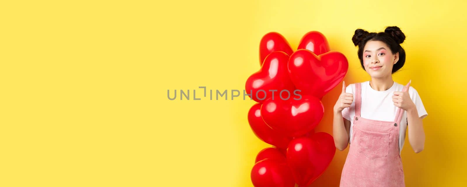 Cute teenage asian girl showing thumbs up, waiting for Valentines day near red heart balloons, wearing outfit for romantic date, yellow background.