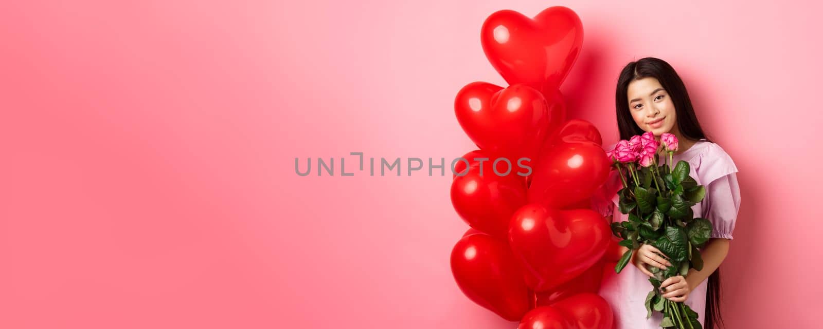 Valentines day concept. Tender and gentle teenage girl holding romantic flowers, standing near red heart balloons from lover and gazing with love, standing on pink background.