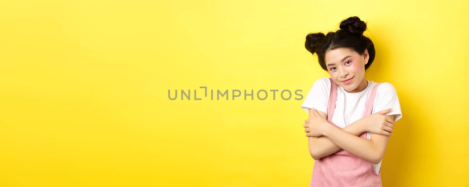 Summer lifestyle concept. Cute asian beauty girl with makeup, hugging herself and smiling romantic, standing tender on yellow background.