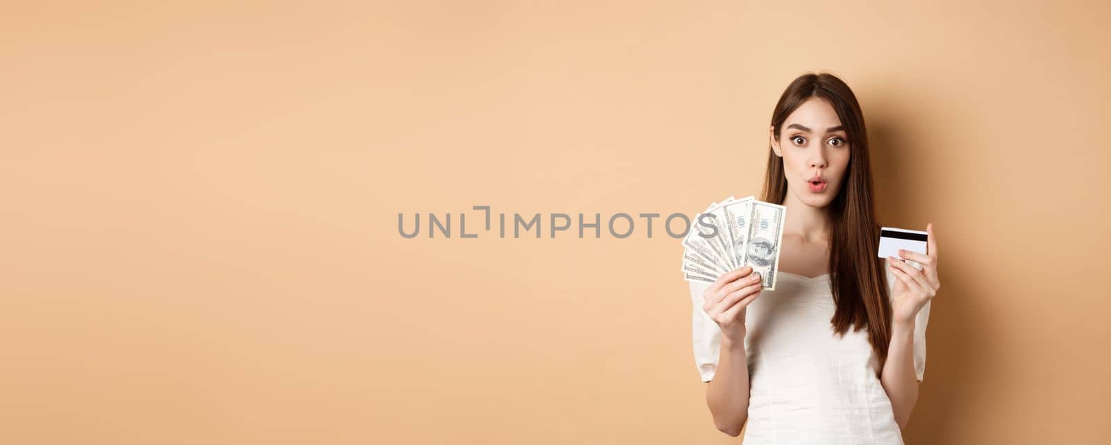 Excited girl showing dollar bills and plastic credit card, saying wow with amazed face, standing on beige background by Benzoix