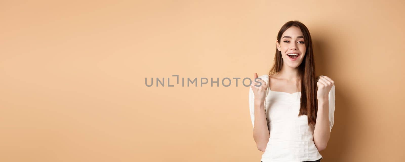 Happy smiling girl celebrating victory, making fist pump and say yes, achieve goal, triumphing or getting motivated, standing on beige background by Benzoix