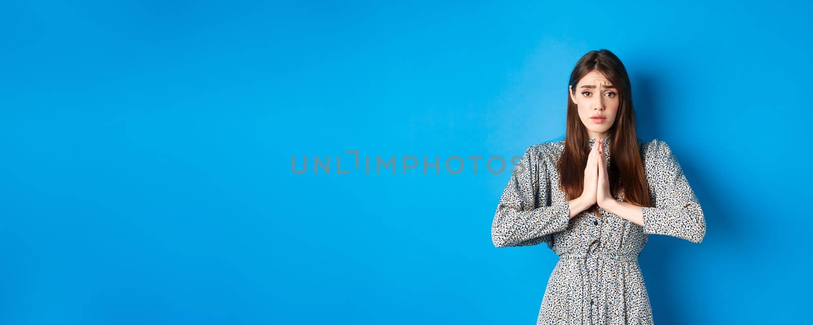 Sad girl asking for help, begging you with troubled face expression, standing in dress on blue background.