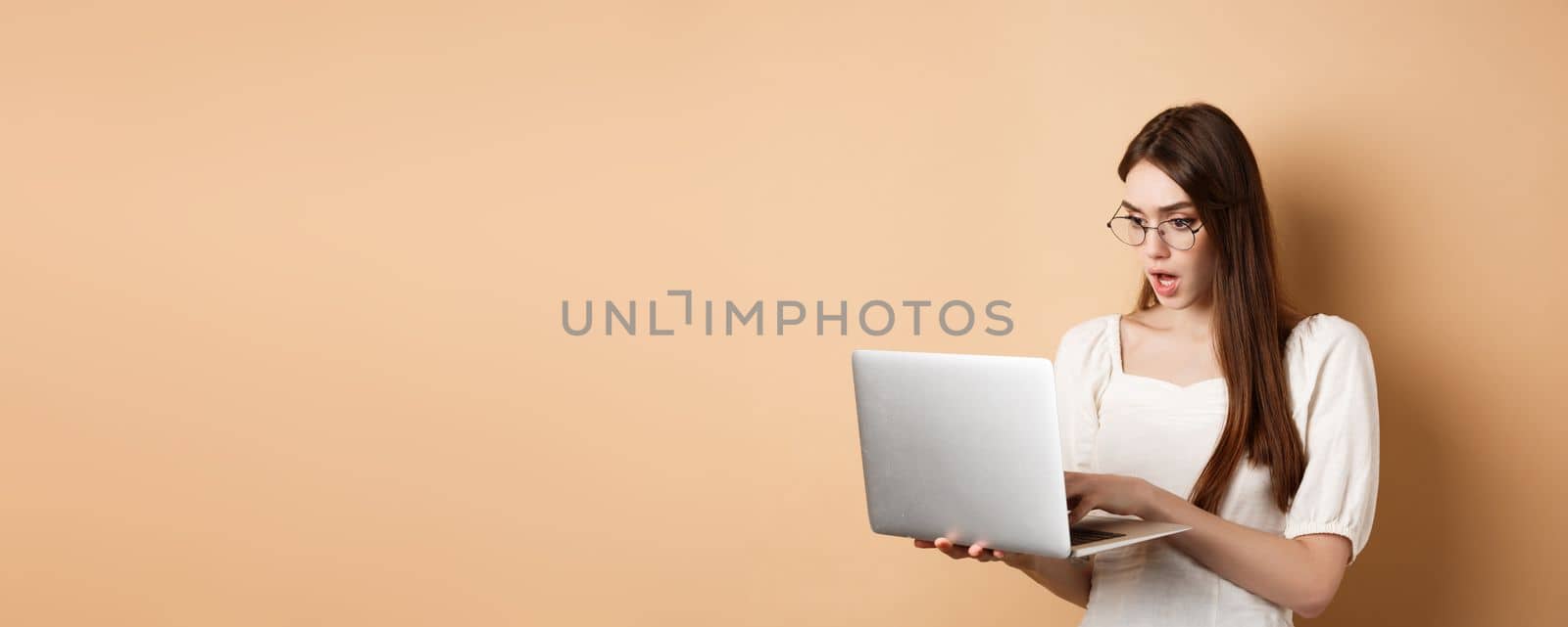 Worried girl look at laptop screen, typing on computer with focused face. Woman freelancer working in internet, standing on beige background by Benzoix