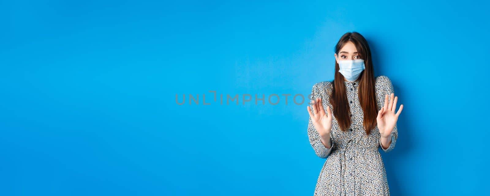 Covid-19, pandemic lifestyle concept. No thank you. Reluctant and worried girl in medical mask asking non-masker to stay away, showing block gesture, tell stop, blue background by Benzoix