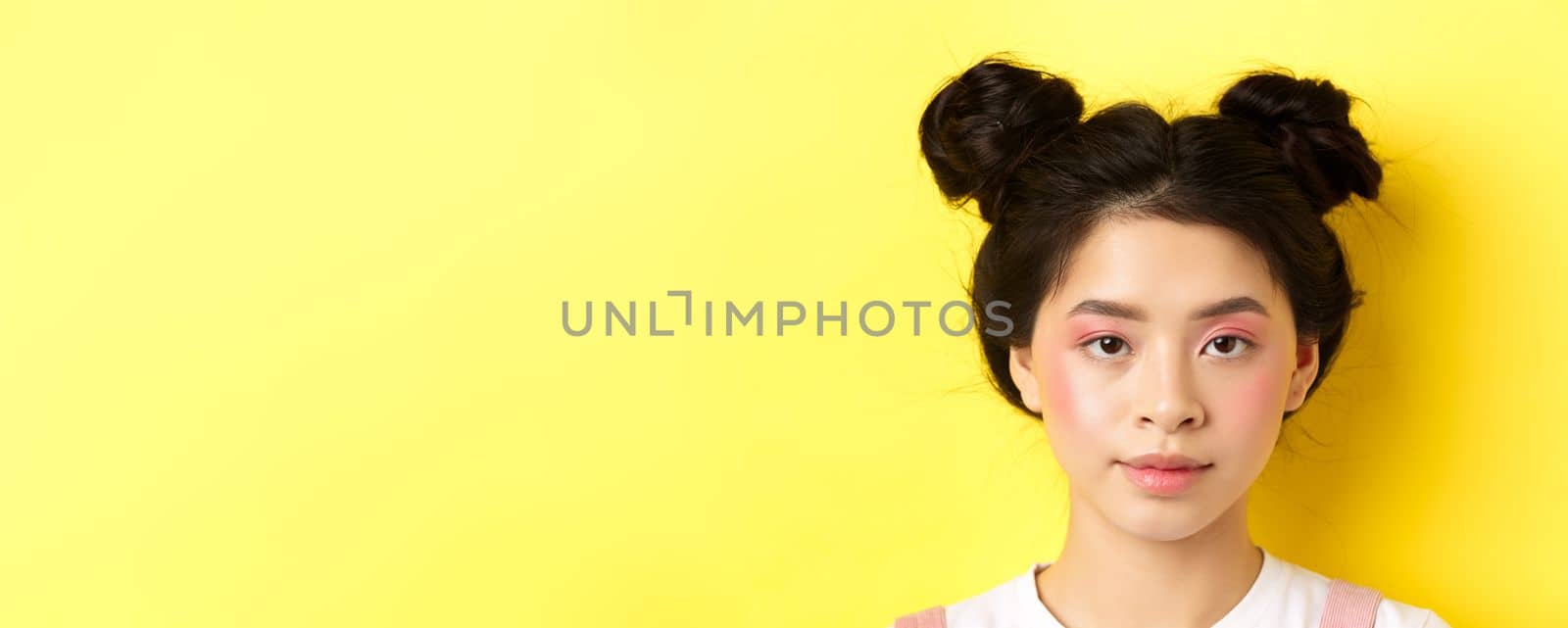 Head portrait of stylish asian girl with bright makeup and hairbuns, looking at camera, standing on yellow background.