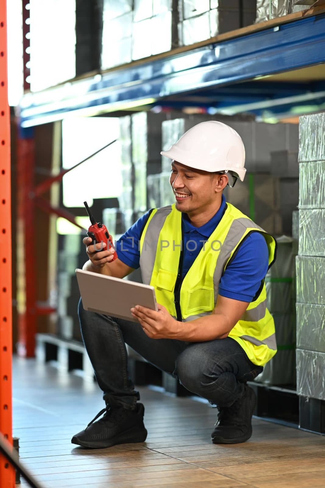 Warehouse worker using digital tablet, checking quantity of storage product in aisle between shelves full of packed boxes.