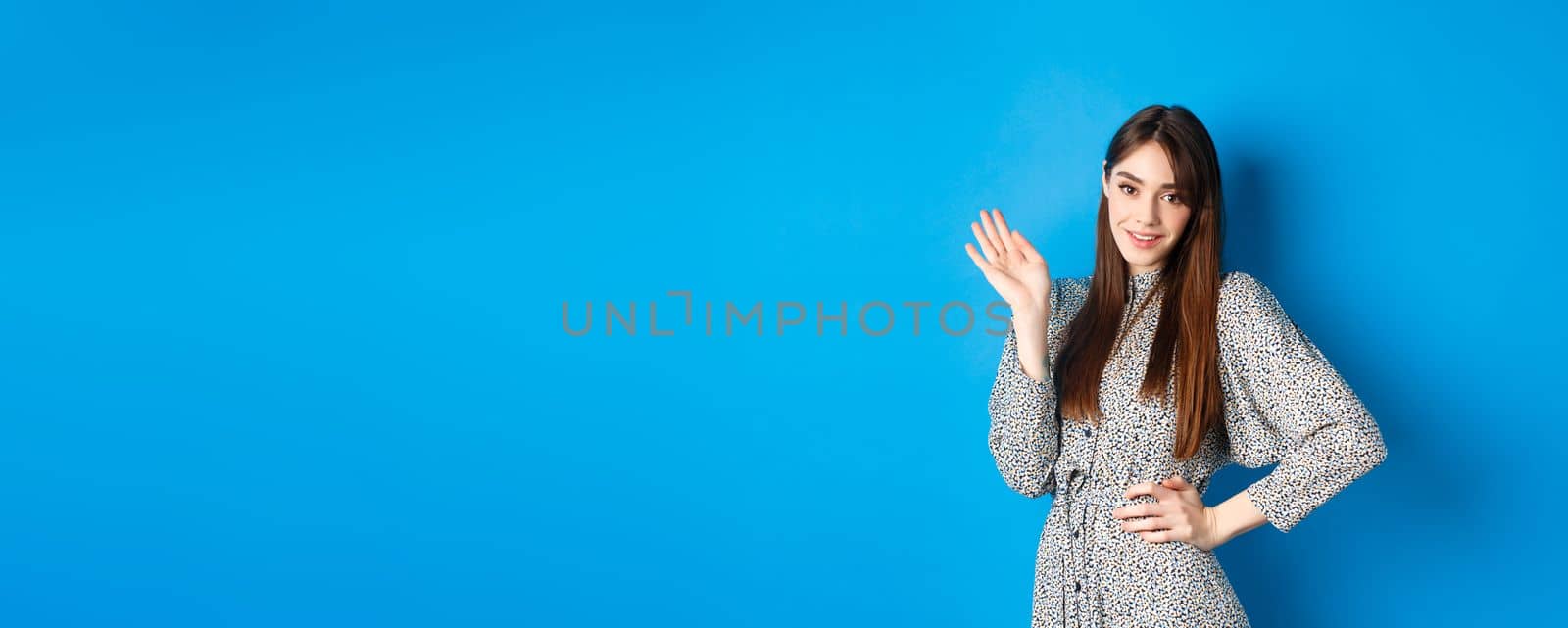 Beautiful lady in dress waving hand and say hello, greeting you with cute smile, standing on blue background by Benzoix