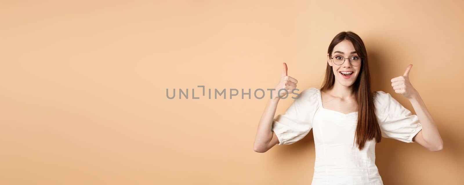 Excited young woman in glasses showing thumbs up and smiling, praising good optic store, approve and like eyewear, standing on beige background by Benzoix