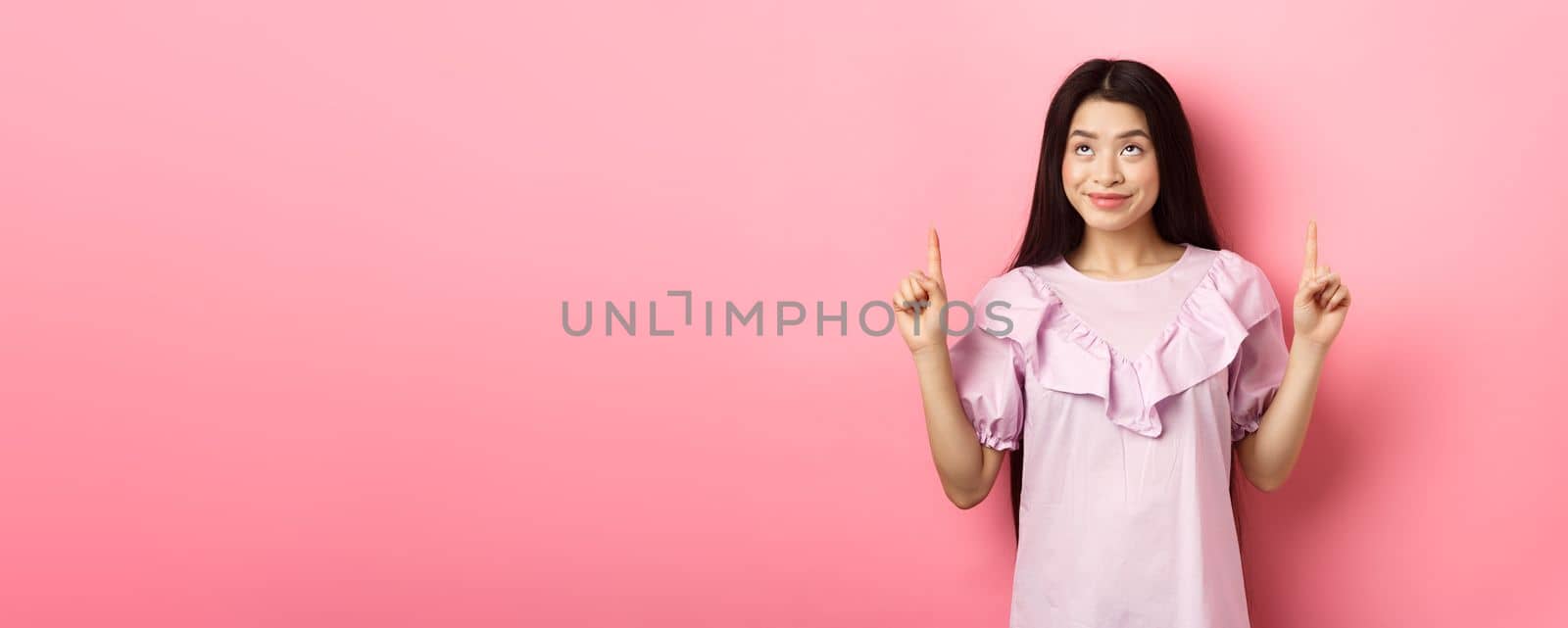 Beautiful smiling asian teen girl pointing fingers up, looking at advertisement with happy face, standing on pink background.