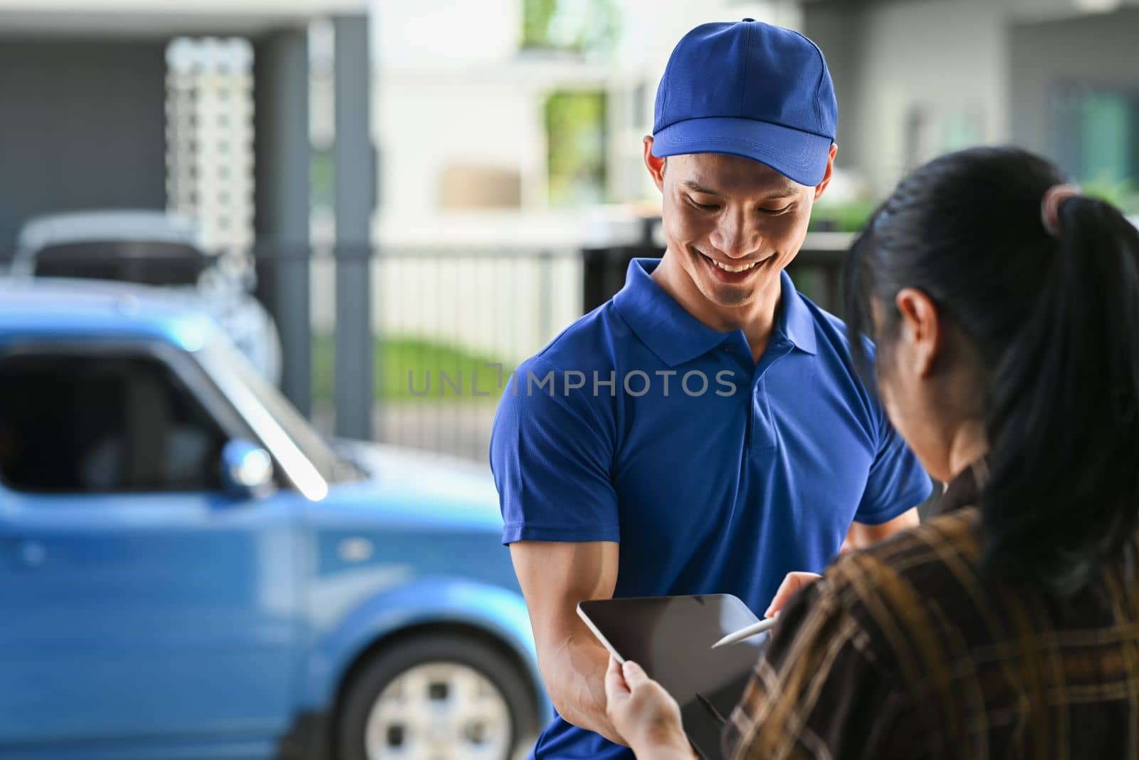 Cropped shot of delivery man in blue uniform using digital tablet while standing at open van full of delivering package by prathanchorruangsak