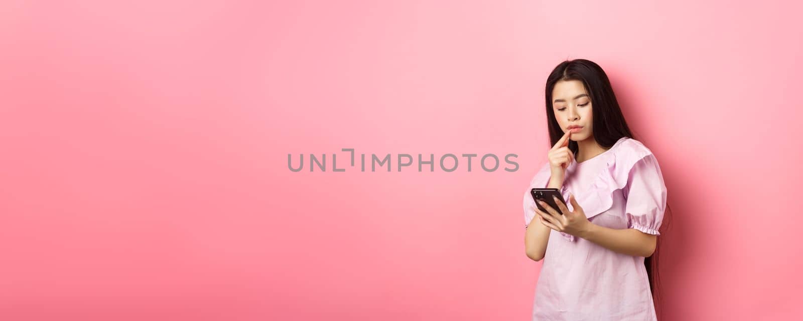 Online shopping. Pensive asian girl looking at smartphone screen, making choice, standing in dress against pink background.