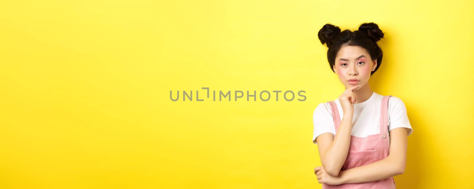 Suspicious asian girl looking at camera and thinking, stare with disbelief, standing with makeup on yellow background.