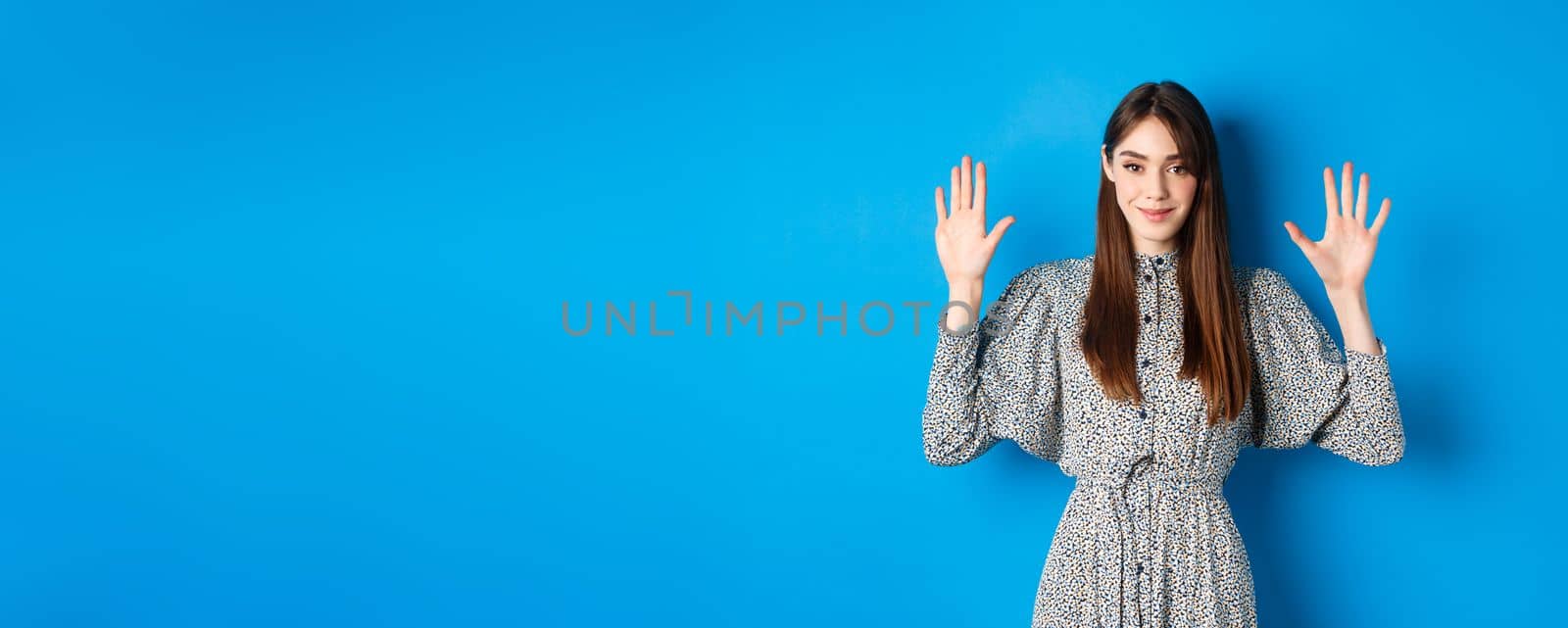 Young beautiful girl in dress with natural long hair, showing number ten with fingers and smiling, standing against blue background by Benzoix