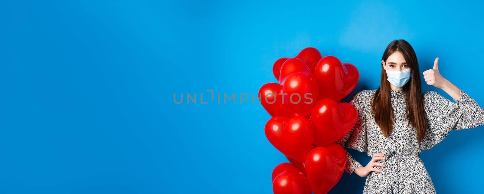 Coronavirus and pandemic concept. Beautiful woman in medical mask and dress standing near Valentines day balloons and showing thumb up, standing on blue background by Benzoix