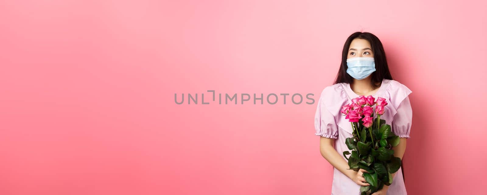 Romantic asian girl in medical mask looking aside at empty space with dreamy eyes, holding bouquet of roses for Valentines day, having date with lover during pandemic.