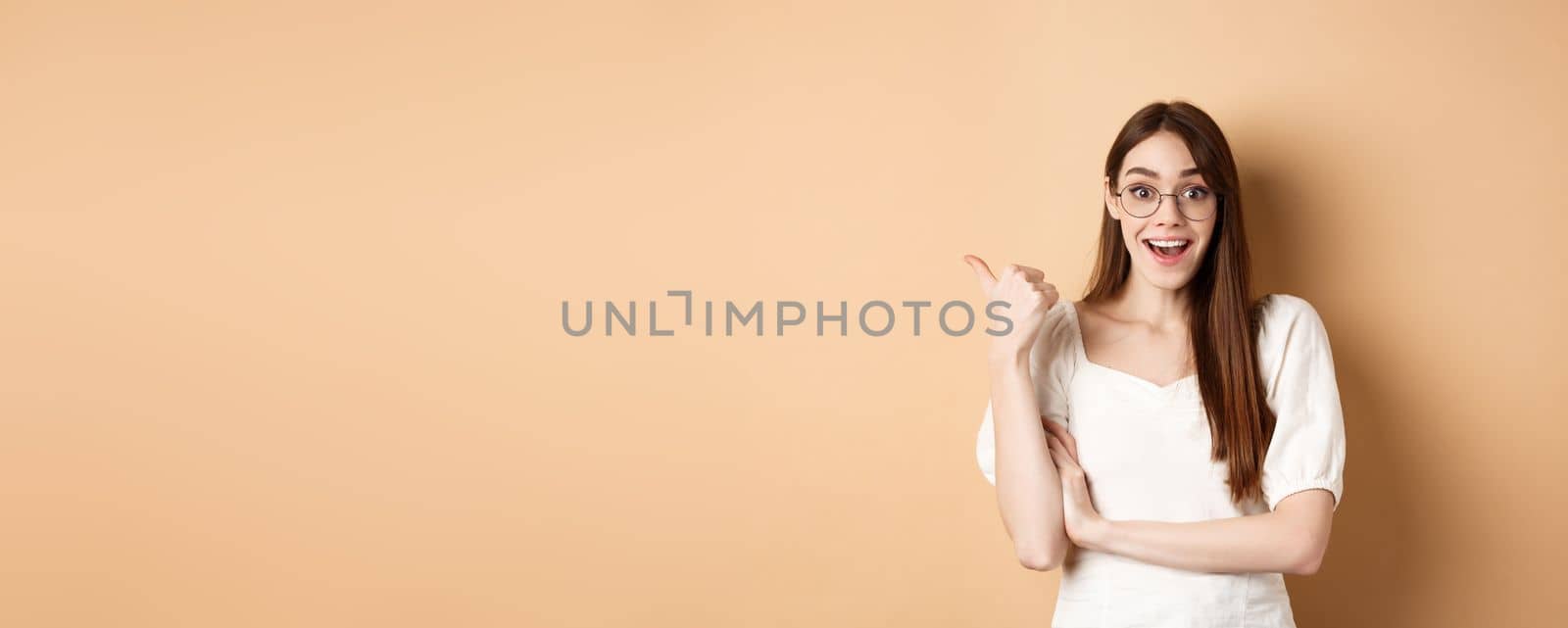 Excited girl in glasses pointing aside at logo, smiling amazed, recommending company, standing on beige background by Benzoix