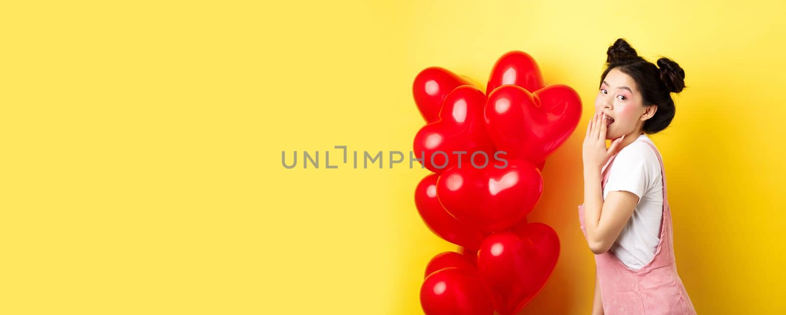 Valentines day and relationship concept. Coquettish and romantic girl laughing, covering mouth with hand, look silly at camera, standing near red heart balloons, yellow background by Benzoix