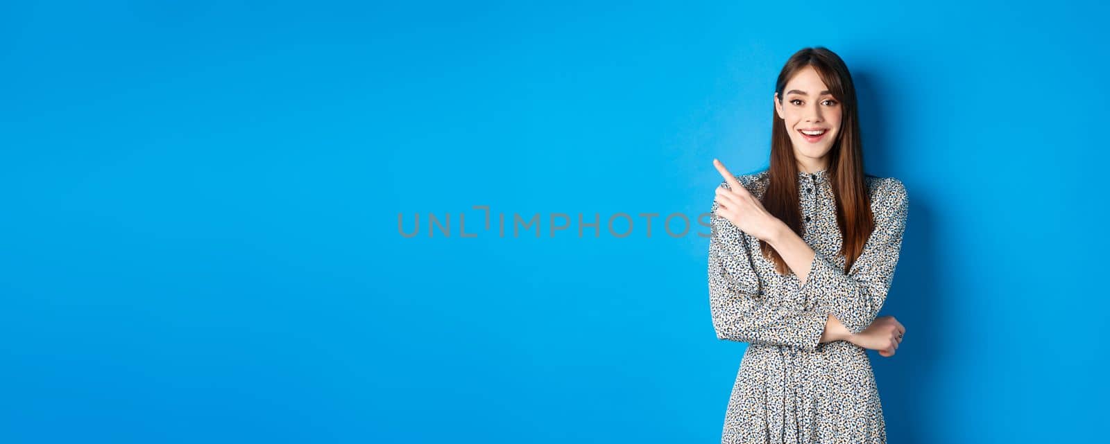 Cheerful young woman in long dress smiling, pointing finger at upper left corner, advertising product on blue background.