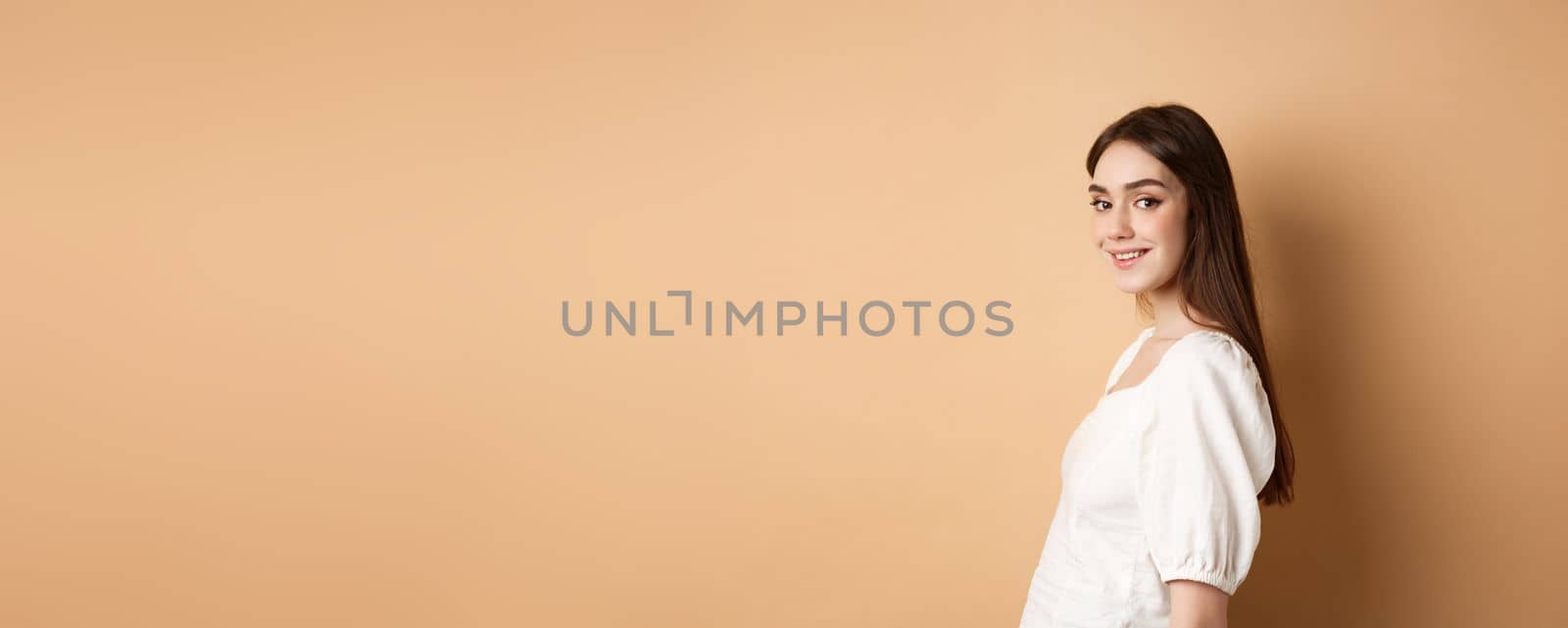 Profile of pretty european girl turn head at camera and smiling coquettish, standing in white blouse against beige background.