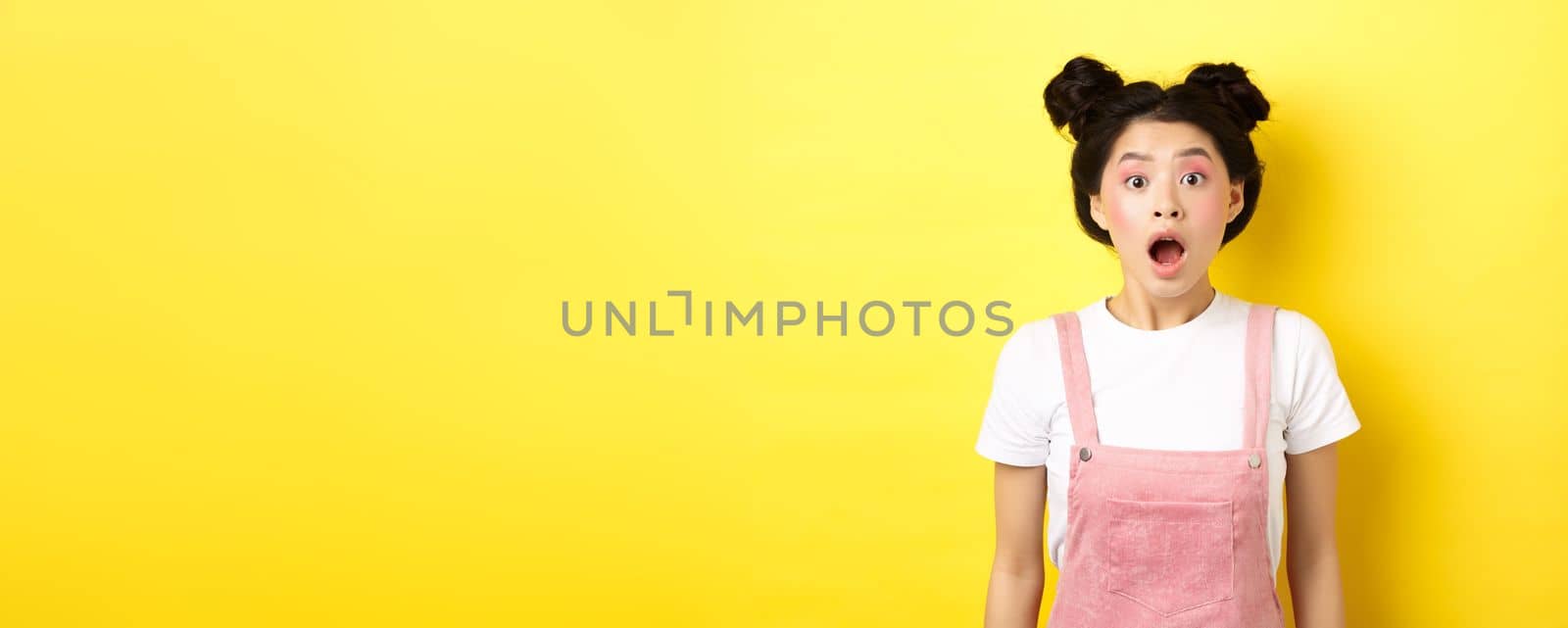 Excited asian teen girl drop jaw, gasping amazed with opened mouth, looking at camera impressed, checking out summer promo offer, standing on yellow background by Benzoix