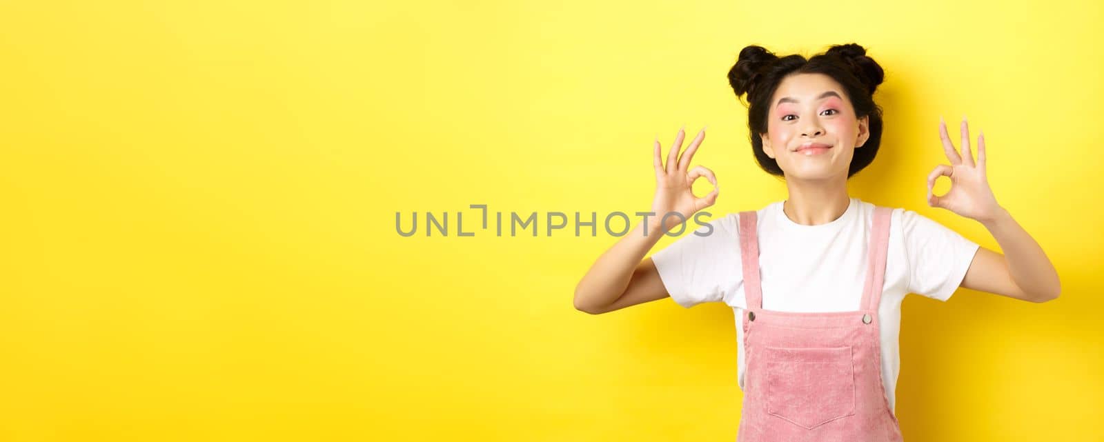 Happy teen koren girl with beauty makeup, showing okay signs and smiling satisfied, recommending product, praise good choice, standing on yellow background.