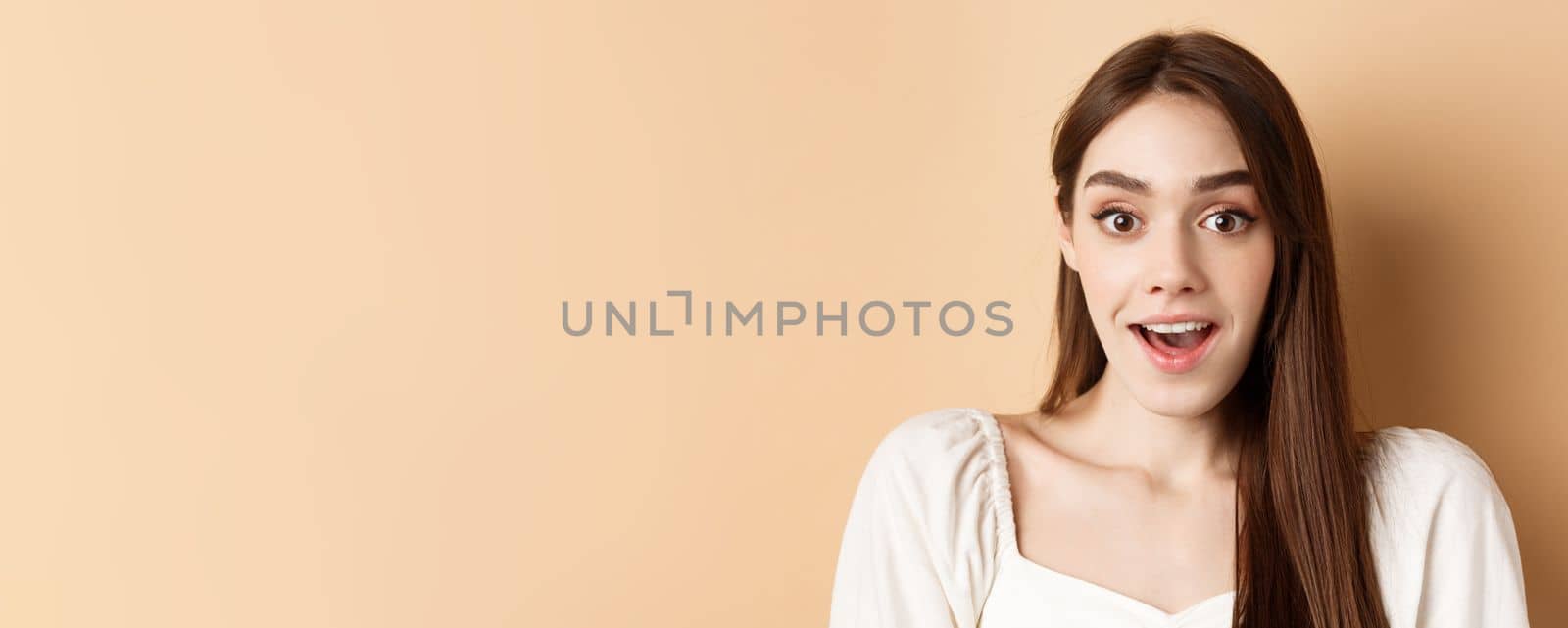 Excited young woman gasping amazed, saying wow and looking at camera, checking out promo offer, standing on beige background.