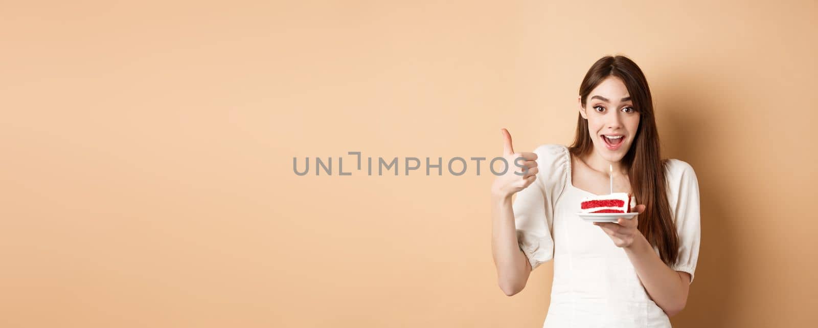 Excited birthday girl show thumb up and hold cake with candle, making wish, satisfied with bday party, smiling happy at camera, beige background by Benzoix